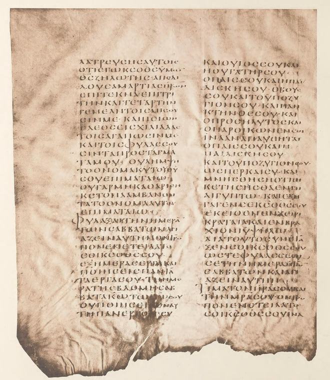 A brown, crinkled parchment leaf with two columns of large Greek capital letters and a torn lower edge.