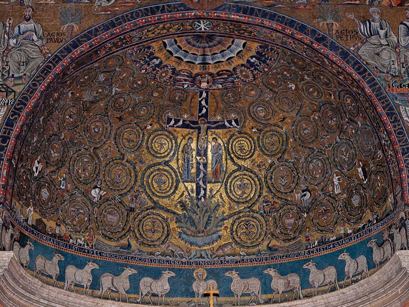 Golden half-dome mosaic showing a dark cross with the crucified Christ rising from an acanthus plant, surrounded by spiraling green and gold vine scrolls, above a blue frieze of white lambs.