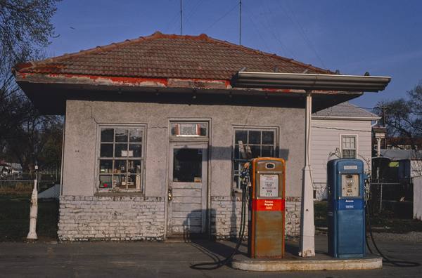 A small stucco filling-station building with a red tile roof and two old gasoline pumps in front, one orange and labeled Amoco Regular, one blue and labeled Amoco lead-free, under a clear blue sky.