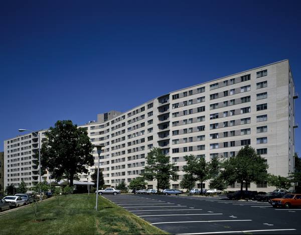 A long, curving multi-story pale-brick apartment building with rows of small windows, trees and a grassy slope in front and a parking lot with parked cars, under a deep blue sky.