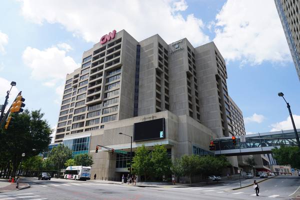 A tall concrete hotel-and-office tower with a red CNN sign on its roof stands over a city street under a blue sky with clouds.