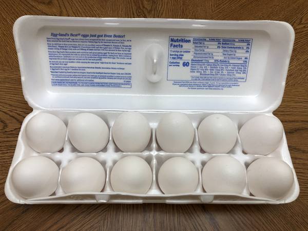 A white foam egg carton lying open on a wooden table, holding twelve white eggs in two rows, with a nutrition label and printed text inside the lid.