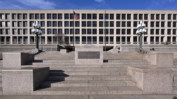 A wide, flat-roofed concrete government building with rows of windows, two flagpoles and two lamp posts, seen from the foot of a broad granite staircase with a plaque on its center block.
