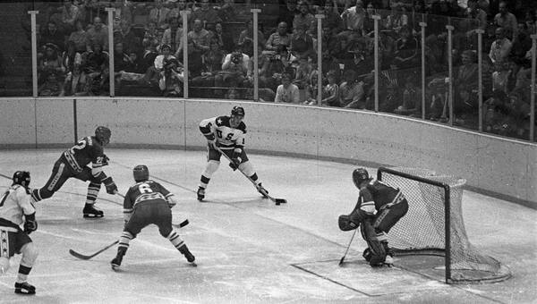 Black-and-white photograph of an ice hockey game: a U.S. player in a white jersey with the puck near the boards, two Soviet players in dark jerseys beside him, the Soviet goalie in his crease at right, and spectators behind the glass.