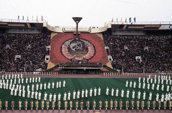 Wide view of a packed stadium during an opening ceremony: rows of performers in white and tan costumes on the green field, and a large red mosaic of the Soviet emblem in the stands beneath the Olympic cauldron.
