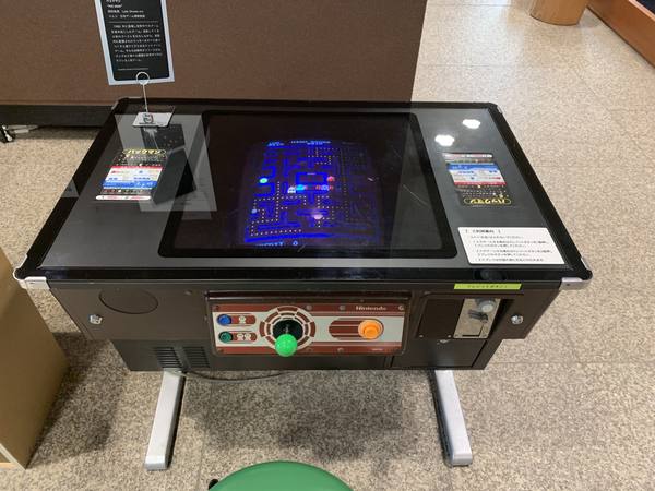 A low black table-style arcade machine seen from above in a museum, its screen showing the blue maze of Pac-Man with small ghosts, a green-topped joystick and buttons on the front edge, and an information placard standing at the upper left.