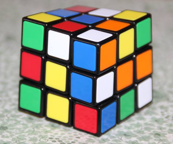 A scrambled Rubik's Cube with a black frame and red, orange, yellow, green, blue and white squares, shown close up on a pale green patterned cloth.