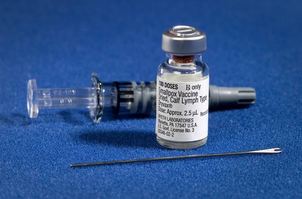 On a blue felt-like cloth, a vial labelled Smallpox Vaccine, Dryvax, stands in front of a syringe lying on its side, with a thin two-pronged bifurcated needle in the foreground.