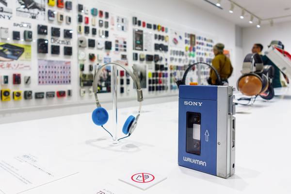 A blue and silver Sony Walkman cassette player standing on a white display table beside a pair of silver headphones with blue foam ear pads, with a wall of other Sony portable players in the blurred background.
