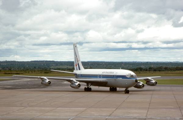 A white Boeing 720 jet with a blue stripe, the words Air Rhodesia on the forward fuselage and a red emblem on the tail fin, standing on a concrete apron under a cloudy sky with low green bush behind.
