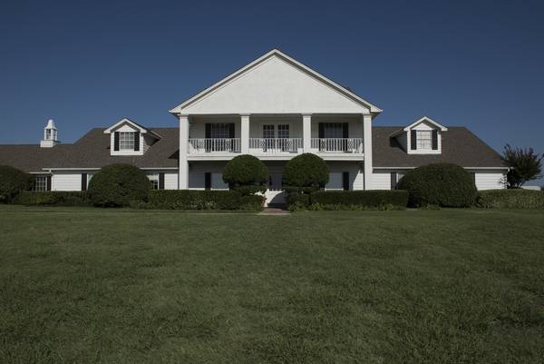 A white house with a tall columned front porch and an upstairs balcony, two dormer windows, round trimmed shrubs along the front and a wide green lawn under a clear dark-blue sky.
