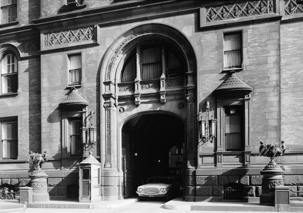 Black-and-white photograph of a tall arched stone-and-brick entrance with ornate carving, wrought-iron lanterns and a small guard booth, with a 1960s car parked inside the archway.