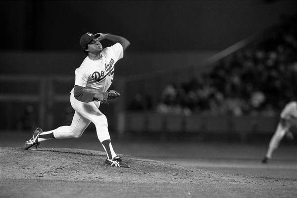A black-and-white night photograph of a Dodgers pitcher in mid-delivery on the mound, his arm cocked back, with the stadium crowd blurred behind him and a second player at the right edge.