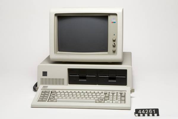 A cream-colored IBM personal computer with a monitor on top of the system unit, two floppy drive slots and a keyboard in front, on a plain background with a black museum number tag at the lower right.