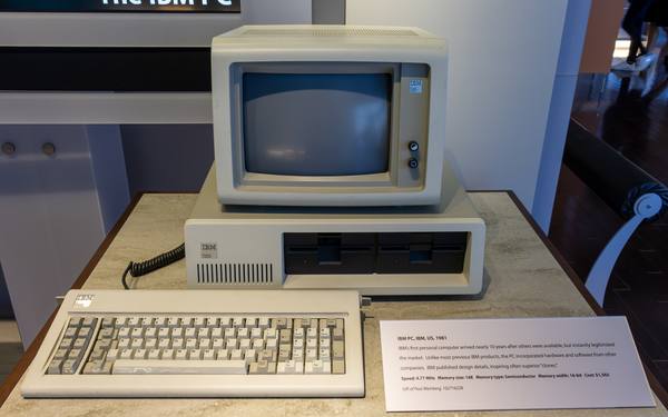 A beige IBM PC with a monitor sitting on the system unit that has two floppy drives, and a separate keyboard in front, on a museum display table with an information label beside it.