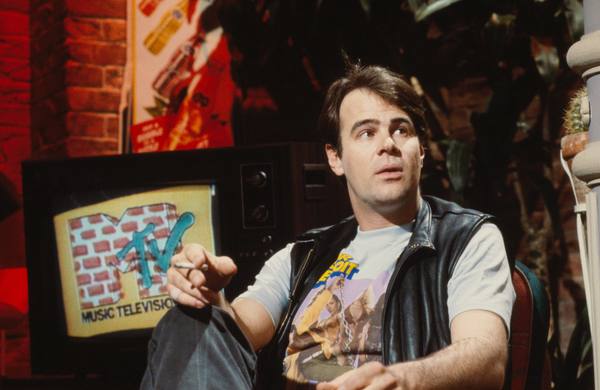A man in a black leather vest and a printed T-shirt sits on a brick-walled television studio set holding a cigarette, with a television beside him showing the brick-pattern MTV Music Television logo.