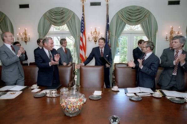 President Reagan standing behind his chair at the Cabinet table, smiling with an arm extended, as men in suits around him applaud, with an American flag behind him and a jar of jelly beans on the table.