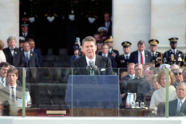 Ronald Reagan in a dark suit speaking at a podium with microphones behind a glass shield, with uniformed military officers and dignitaries standing behind him and seated guests at right.