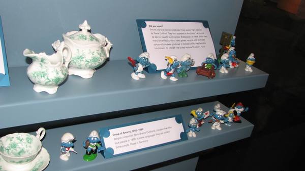 Two blue shelves in a museum case holding small blue Smurf figurines in white caps in various poses, next to green-patterned white china, with two printed museum labels about the Smurfs.