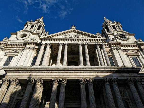 Looking up at the classical stone west front of St Paul's Cathedral, with paired columns, a sculpted pediment and two towers, one with a clock, against a deep blue sky.