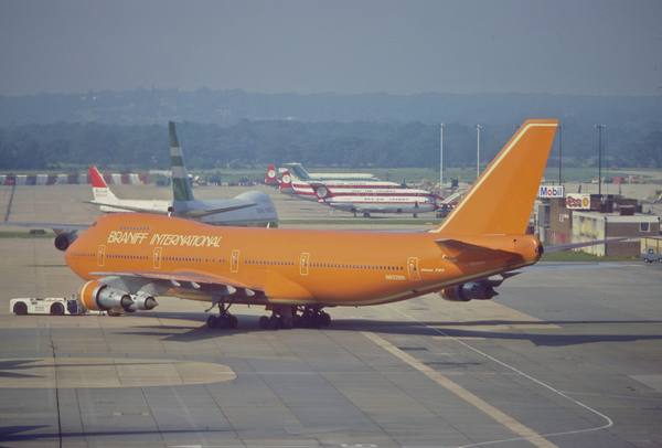 A bright orange Boeing 747 with the words Braniff International on its fuselage and the registration N602BN, standing on an airport apron with a small tug at its nose and other airliners and airport buildings in the hazy background.