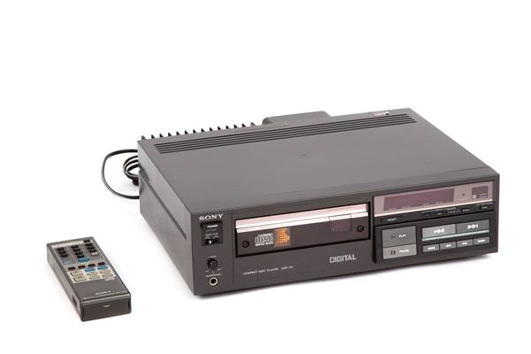 A black Sony CDP-101 compact disc player with a disc tray and a red display, and a small remote control beside it, on a white background.