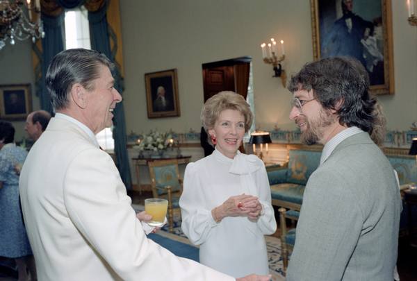 Ronald Reagan in a white dinner jacket, Nancy Reagan in a white blouse and Steven Spielberg, bearded and in glasses and a grey jacket, chatting and smiling in the blue-and-gold Blue Room of the White House.