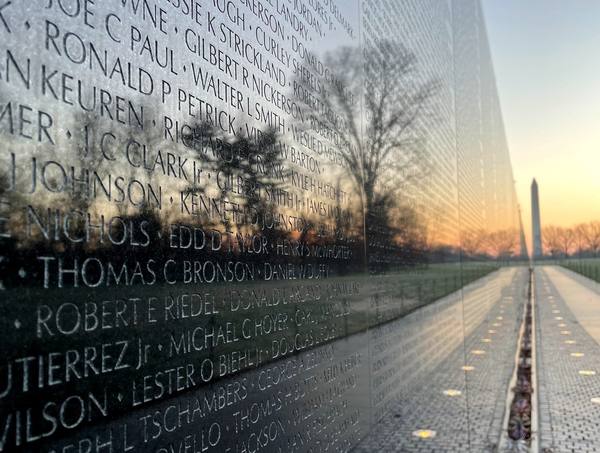 Close, angled view of rows of engraved names on the polished black granite wall, reflecting bare trees against a glowing sky, with the Washington Monument far down the path.