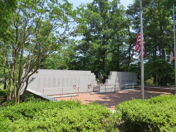 A pale stone memorial wall with rows of engraved names and the words 'They came in peace', a small bronze figure at its center and wreaths on the wall, framed by green trees and a U.S. flag at half-staff.