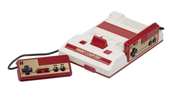 A red and white Famicom game console with one controller docked at its side and the other lying in front on its cord, on a white background.