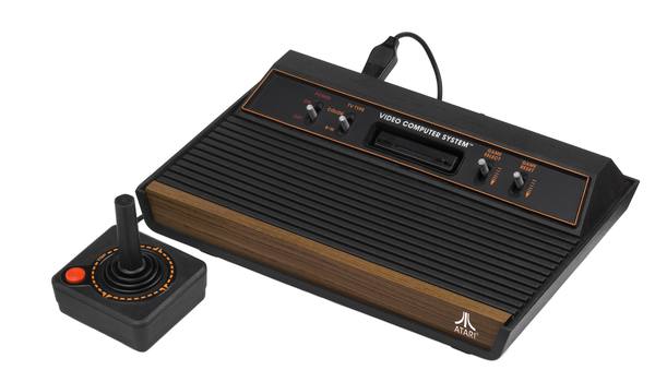 A black Atari game console with a woodgrain front panel, a ridged top, four switches and the words Video Computer System and Atari, beside a black joystick controller with an orange fire button, on a white background.