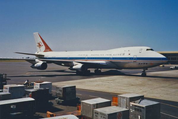 A white Korean Air Lines Boeing 747 with a blue stripe and a red tail on an airport apron, with baggage containers in the foreground.