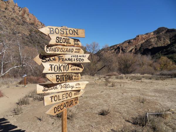 A tall wooden post with a stack of hand-painted arrow signs pointing to distant cities, standing in a dry canyon with rust-colored rock hills under a clear blue sky.