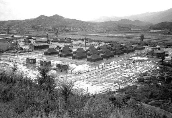 Black-and-white view from a hillside over a valley in Korea showing rows of large army tents and a few huts enclosed by wire fencing on muddy, puddled ground, with hills and mountains in the distance.