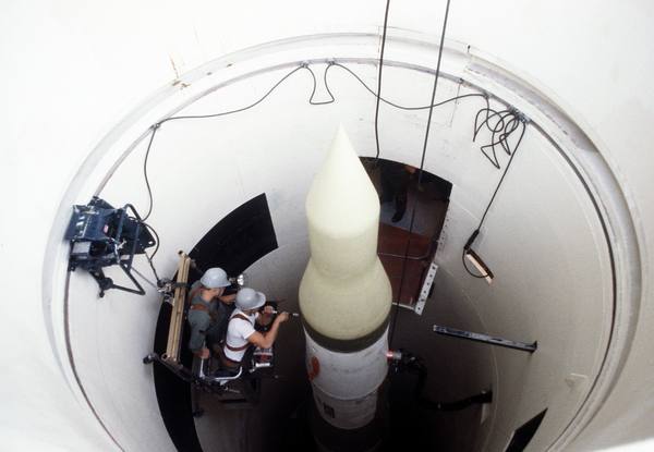 A view looking straight down a missile silo at the pale nose cone of a Minuteman missile, with two crewmen in grey hard hats working on a platform beside it.
