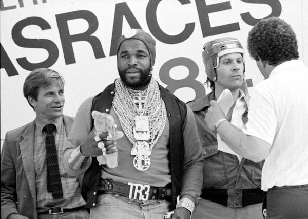 A black-and-white photograph of a man wearing many gold chains, a cap and a belt buckle reading TR3, holding a small plush toy, with a man in a jacket and tie on his left, a man in a helmet and jacket on his right, and a man seen from behind at the edge.