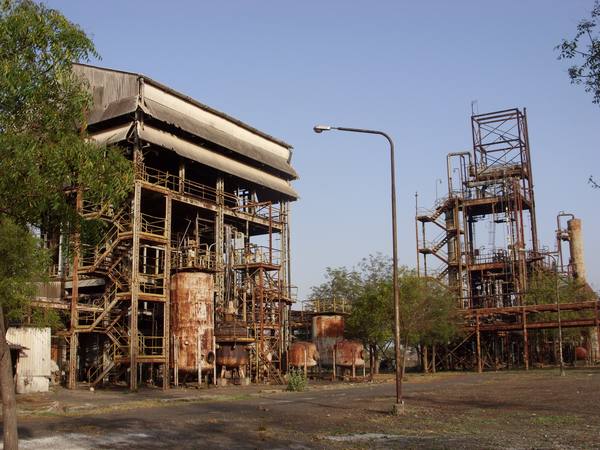 Rusted steel process towers, tanks and staircases of an abandoned chemical plant stand beside an empty road and a street lamp under a pale blue sky.