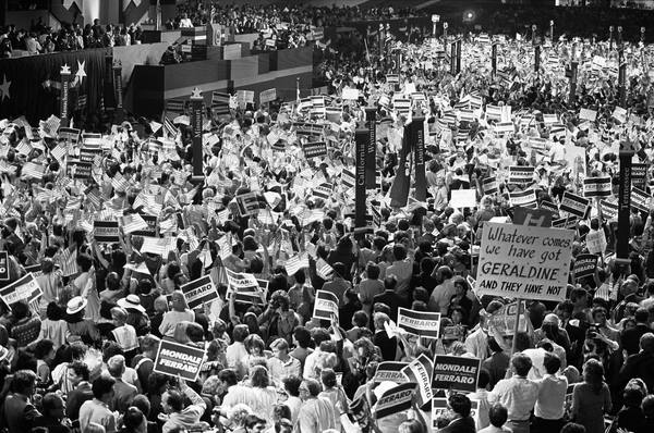 Black-and-white view over a packed convention floor, with delegates waving small American flags and holding Mondale-Ferraro signs, state standards on tall poles, a handwritten sign reading 'Whatever comes, we have got Geraldine and they have not', and a stage podium at the upper left.