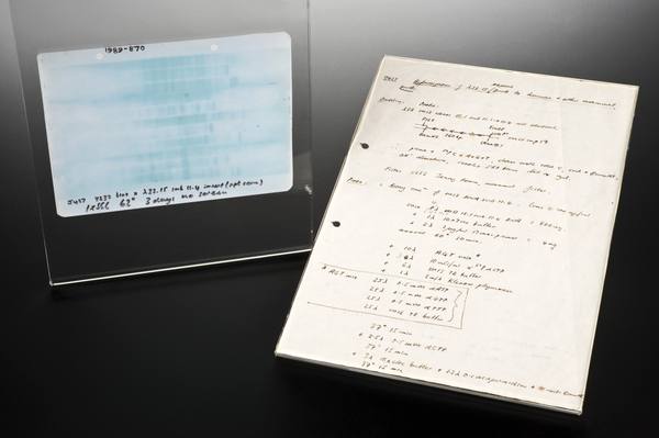 A pale blue film with faint dark bands in a clear holder, marked 1989-870, beside a handwritten laboratory notes page, on a dark glossy surface.