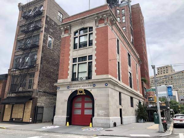A red-brick and limestone firehouse with a large red arched door and the words Hook & Ladder 8 carved above it, seen from a street corner, with a black fire escape on the neighboring building and a white car at the right edge.