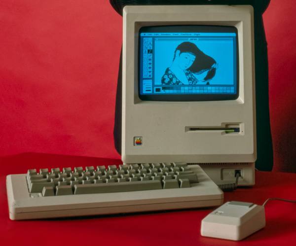A beige all-in-one Macintosh computer with a blue-tinted screen showing a drawing of a woman combing her hair in a drawing program window, with a separate keyboard and a mouse in front of it against a red background.