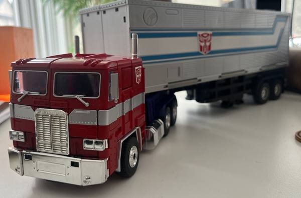 A red and silver toy semi-truck cab pulling a white and silver trailer marked with a red Autobot emblem and blue stripes, sitting on a pale desk by a window, with the trailer softly out of focus.