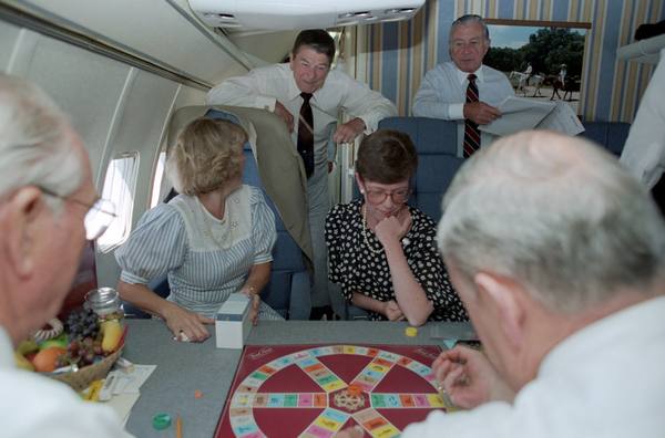 Inside an aircraft cabin, people sit around a table with a round red Trivial Pursuit board and a box of cards, while President Reagan and another man lean over the seat backs behind them and two other players are seen from behind in the foreground.