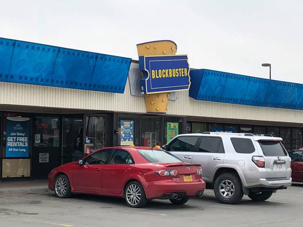 The front of a one-storey shopping-centre store with a blue film-strip roofline and a yellow and blue ticket-shaped Blockbuster sign, with a red sedan and a silver SUV parked in front.