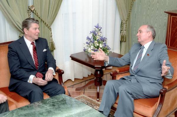 Mikhail Gorbachev, seated at right in a gray suit and gesturing with open hands, talks with President Ronald Reagan, seated at left in a dark pinstripe suit, in a room with green curtains and a vase of flowers.