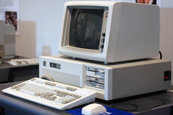 A cream-colored IBM Personal Computer/AT with a dark, switched-off monitor on top, two floppy disk drives on the front, a beige keyboard in front and a white mouse at the bottom edge.