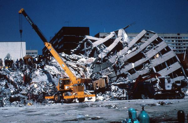 A tilted, partly collapsed concrete apartment block with a yellow crane and an excavator at its base, rubble in front and a deep blue sky behind.