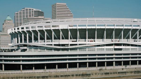 The white, ring-shaped concrete exterior of Riverfront Stadium beneath office towers of downtown Cincinnati on a clear day.