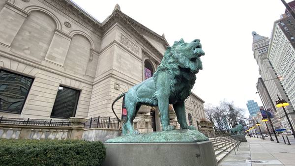 A weathered green bronze lion statue on a stone pedestal stands in front of the stone facade of the Art Institute of Chicago, with tall buildings lining the street on the right.