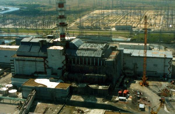 Aerial view of the grey concrete sarcophagus over Chernobyl's reactor 4 with a tall red-and-white ventilation stack, an orange crane beside it, and switchyard pylons beyond.